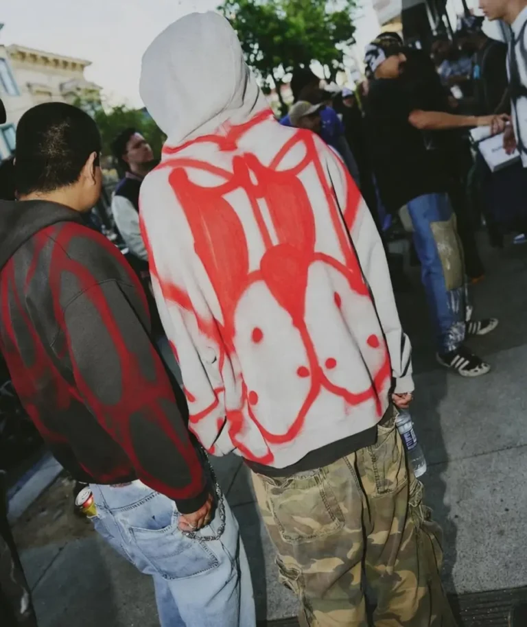 Rear-view street-style photograph of two attendees outside the SVRN San Francisco flagship during the Chito x SVRN S/S26 launch event, featuring oversized hoodies with bold red airbrushed graffiti graphics, camouflage cargo pants, distressed denim, and a crowd gathered along Fillmore Street