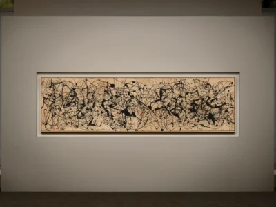Wide installation view of Jackson Pollock’s Number 7A, 1948, mounted on a minimalist gallery wall. The monumental horizontal canvas features Pollock’s signature Abstract Expressionist drip technique, with energetic black enamel lines layered across raw beige canvas and punctuated by scattered red accents, creating a dense web of movement, rhythm, and controlled chaos