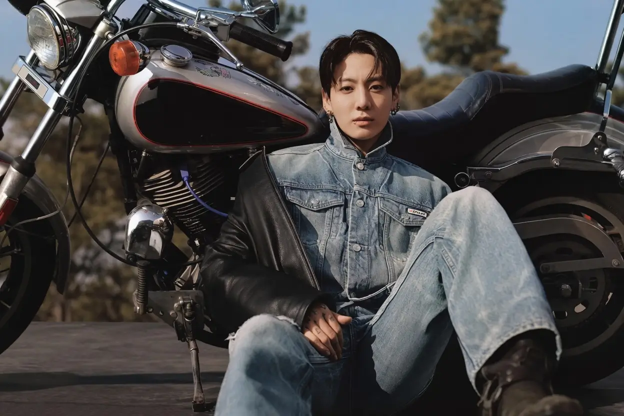 Lifestyle campaign image of a young man seated beside a classic motorcycle wearing a light-wash denim set layered with a black leather jacket. The look combines vintage Americana and biker influences, with faded denim textures, relaxed proportions, and a cinematic outdoor backdrop emphasizing rugged, effortless style