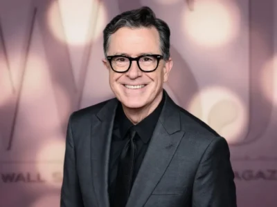 Portrait of a smiling man wearing black-framed glasses, a charcoal gray suit, black shirt, and black tie while posing in front of a softly blurred event backdrop with warm circular lighting and muted pink-gray tones