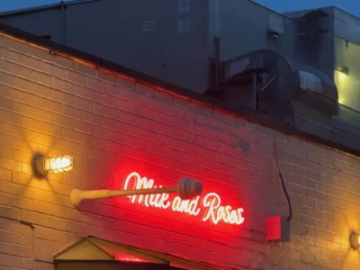 Exterior view of Milk and Roses in Greenpoint, Brooklyn at dusk, featuring the restaurant’s glowing red neon sign mounted on a painted brick façade, illuminated by warm industrial wall lights against a deep blue evening sky