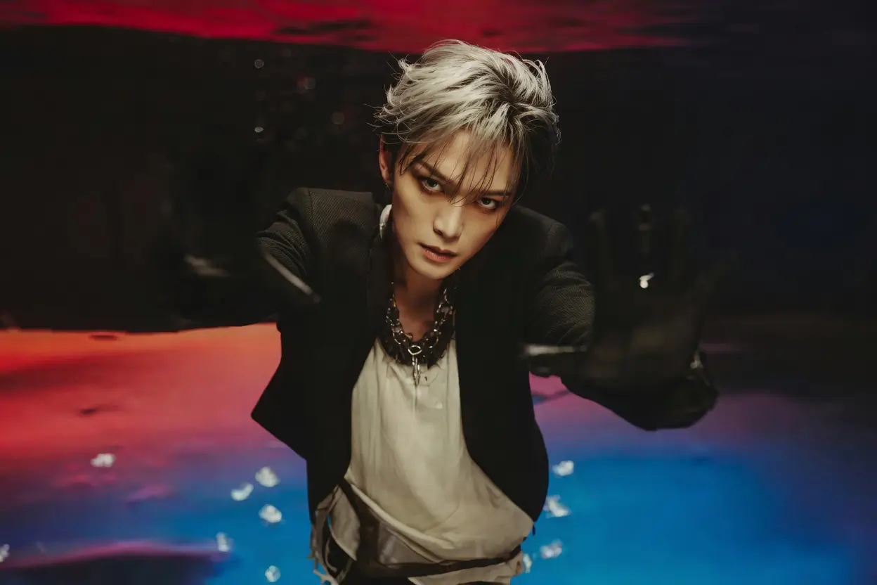 Cinematic portrait of Kim Jaejoong leaning toward the camera in a dark, moody setting illuminated by red and blue lighting. He wears a black tailored jacket layered over a light-colored shirt with a silver chain necklace. His silver-gray hair is styled in a tousled, textured look that frames his face, creating a sharp and dramatic silhouette. The composition carries a sleek K-pop visual identity with a high-fashion editorial atmosphere and a slightly rebellious edge