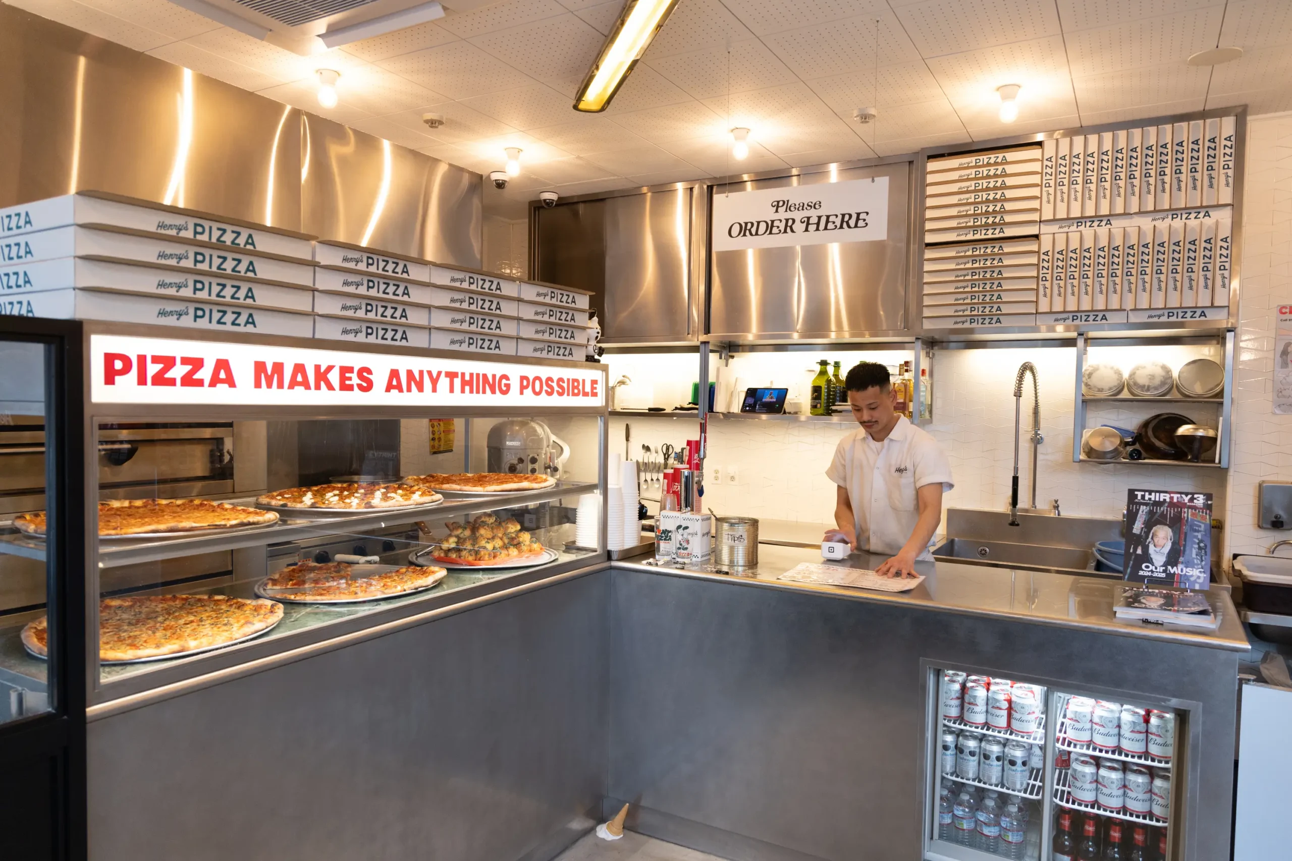 Wide interior view of a contemporary New York-style pizza shop featuring an open kitchen and industrial stainless-steel finishes. Stacks of branded pizza boxes line the upper shelves while a bold illuminated sign above the display case reads, “PIZZA MAKES ANYTHING POSSIBLE.” Multiple whole pizzas and baked items are showcased behind glass at the counter. A staff member in a white shop shirt prepares orders behind the register beneath a hanging “Please ORDER HERE” sign. Warm overhead lighting, metallic surfaces, and neatly organized shelving create a clean yet creative atmosphere that blends neighborhood pizzeria energy with a design-forward lifestyle aesthetic