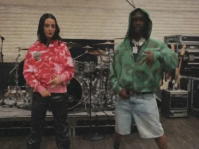 Grainy, lo-fi photo of Katy Perry and Chief Keef in a music studio. Katy in pink camo hoodie on the left, Chief Keef in green camo hoodie pointing forward on the right, surrounded by drums and equipment