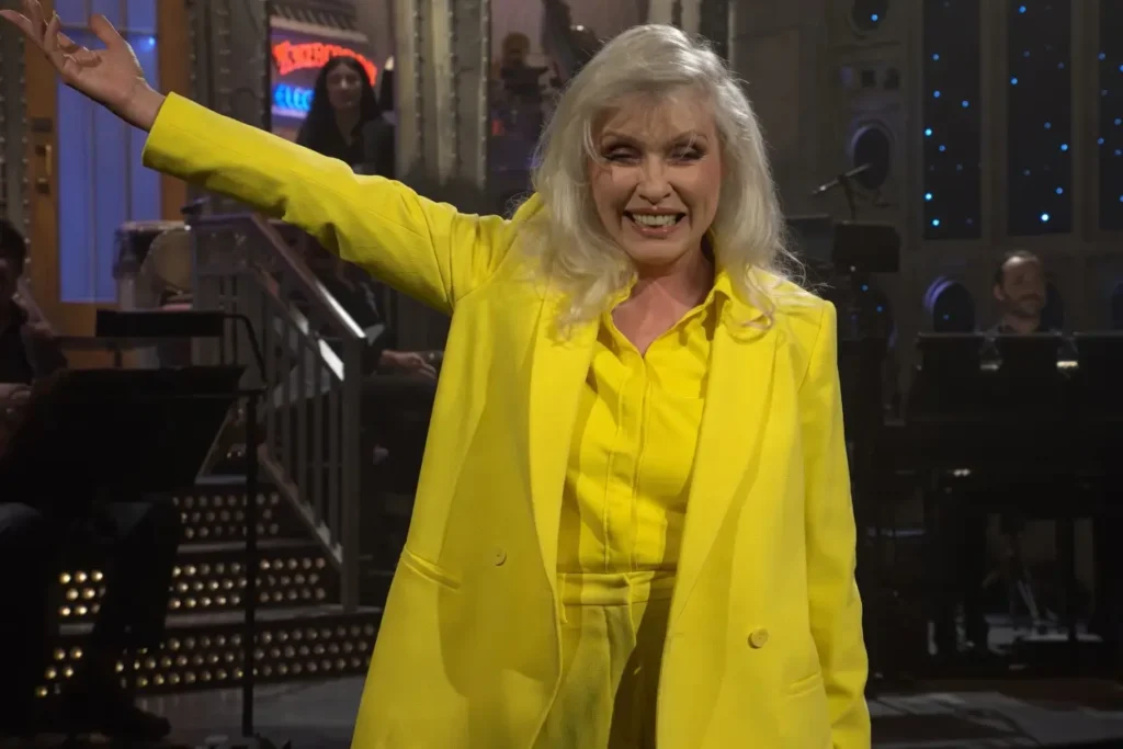 Debbie Harry wearing a bright yellow blazer on the Saturday Night Live stage, raising her arm while introducing Olivia Rodrigo during a live performance moment