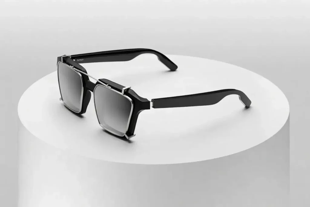 A pair of structured black sunglasses rests on a cylindrical white pedestal, presented in a clean, studio-lit environment. The frames combine sharp, geometric lines with a slightly industrial edge, featuring a dual-material look—matte black acetate contrasted by a thin metallic bar that runs across the top and connects the lenses. The square lenses carry a soft gradient tint, adding depth while maintaining a sleek, understated tone. The temples extend in a sculpted, angular form, reinforcing the design’s modern, almost architectural character. Overall, the composition emphasizes precision and craftsmanship, with a quiet balance between technical detailing and minimalist refinement