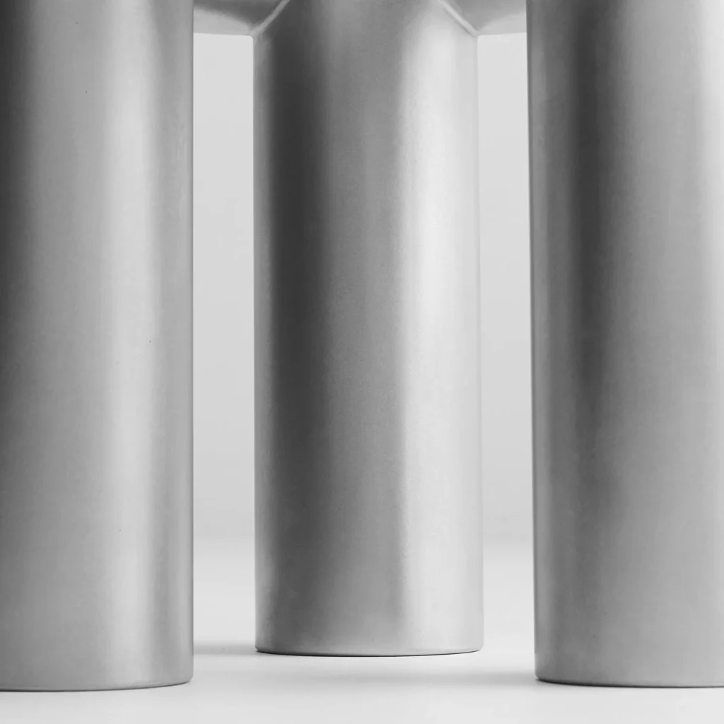 A close-up studio photograph of three oversized brushed stainless steel cylindrical legs supporting a sculptural furniture piece. The matte metallic finish and soft monochrome lighting emphasize the object’s industrial texture, seamless construction, and minimalist collectible design aesthetic
