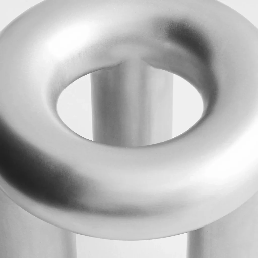 A close-up photograph of a sculptural brushed stainless steel stool featuring a hollow donut-shaped seat with softly rounded contours. The matte metallic surface diffuses light across the curved form, while the cylindrical legs visible beneath reinforce the piece’s minimalist industrial and collectible design aesthetic