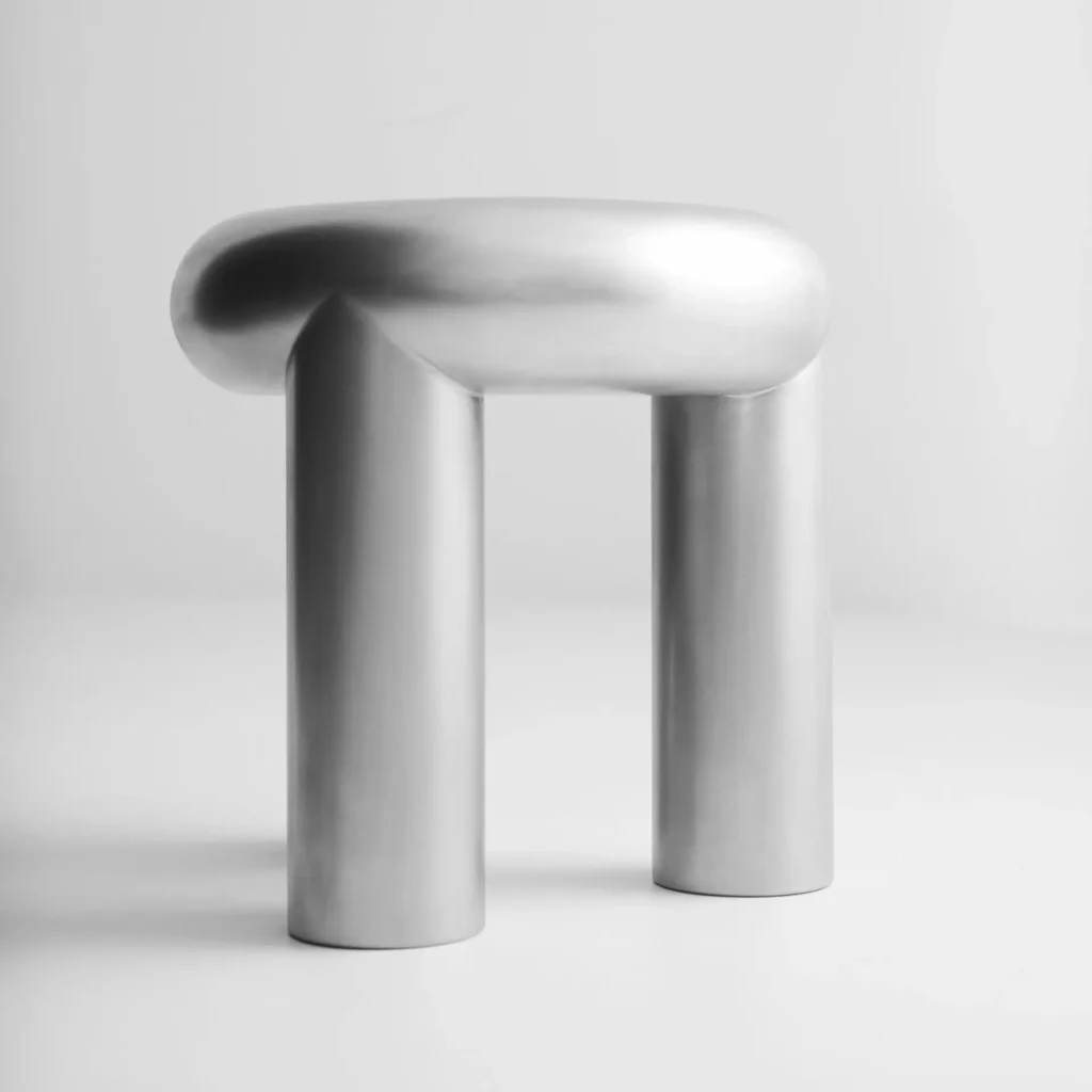 A minimalist sculptural stool crafted from brushed stainless steel, featuring a rounded pill-shaped seat supported by two oversized cylindrical legs. Set against a monochrome studio backdrop, the collectible design piece highlights soft industrial curves, seamless metalwork, and a muted matte finish that subtly captures light