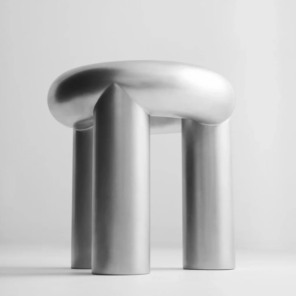 A sculptural brushed stainless steel stool with a rounded organic seat balanced on three oversized cylindrical legs. Photographed in a minimalist monochrome studio setting, the collectible furniture piece emphasizes soft industrial curves, seamless construction, and a tactile matte metallic surface