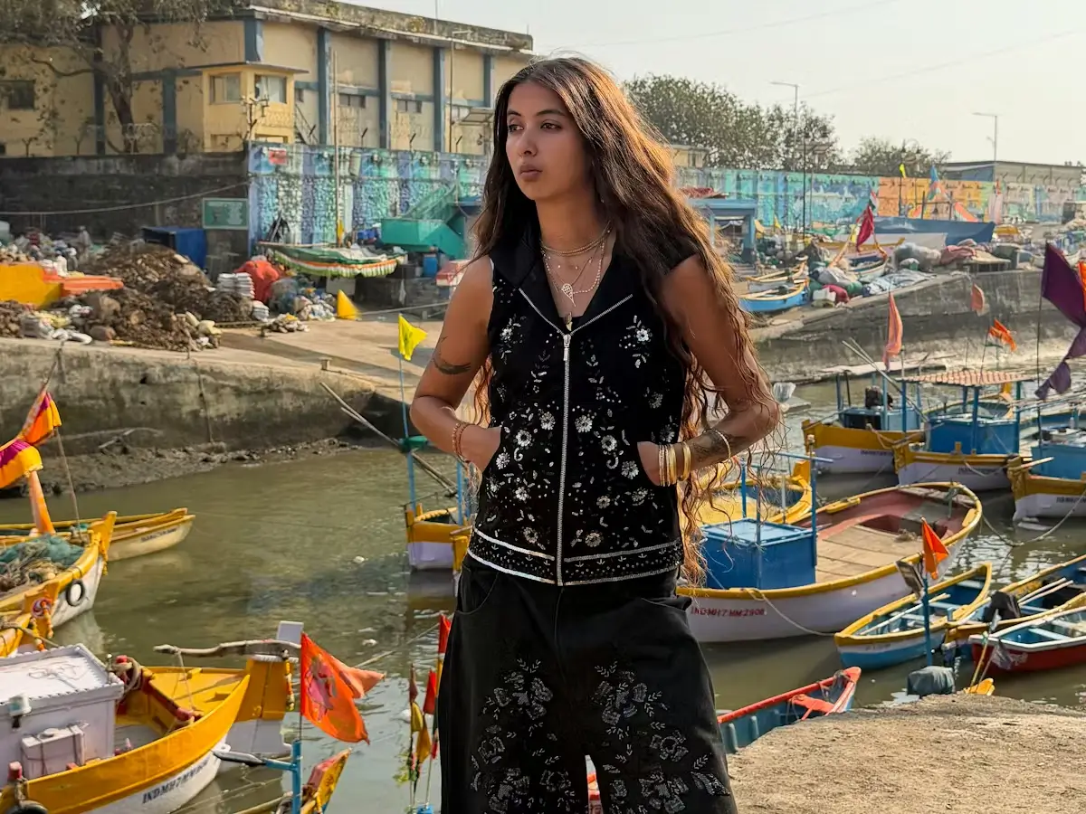 A woman wearing a black embroidered sleeveless zip vest and matching wide-leg trousers stands beside a crowded fishing harbor, layered with gold jewelry as colorful boats and weathered waterfront buildings frame the sunlit coastal backdrop
