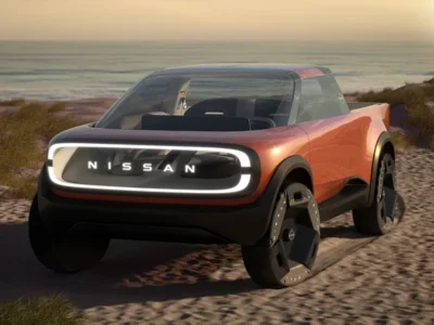 Front three-quarter view of the Nissan Surf-Out Concept electric pickup parked on coastal sand dunes at sunset, featuring a transparent illuminated grille, glowing perimeter light signature, oversized aero wheels, and a minimalist two-door cabin