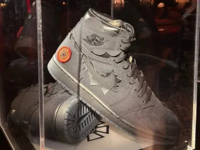 A display-case presentation of a black high-top Air Jordan shoe showcased under warm ambient lighting inside a transparent acrylic box. The shoe features a rugged canvas-textured upper with tonal layered overlays, jagged deconstructed paneling, and a fragmented black Swoosh integrated into the sidewall. Embossed Air Jordan Wings branding appears near the collar, while an orange circular heel patch with a dark Jumpman-style emblem introduces a subtle contrast against the monochrome charcoal palette. The matte black midsole and smoky translucent outsole reinforce the stealth aesthetic, with a second sneaker partially visible beneath to expose the outsole traction pattern. Reflections on the acrylic case and the dimly lit lounge-like environment in the background create a luxury showroom atmosphere, emphasizing the shoe collectible, sample-like presentation and experimental craftsmanship