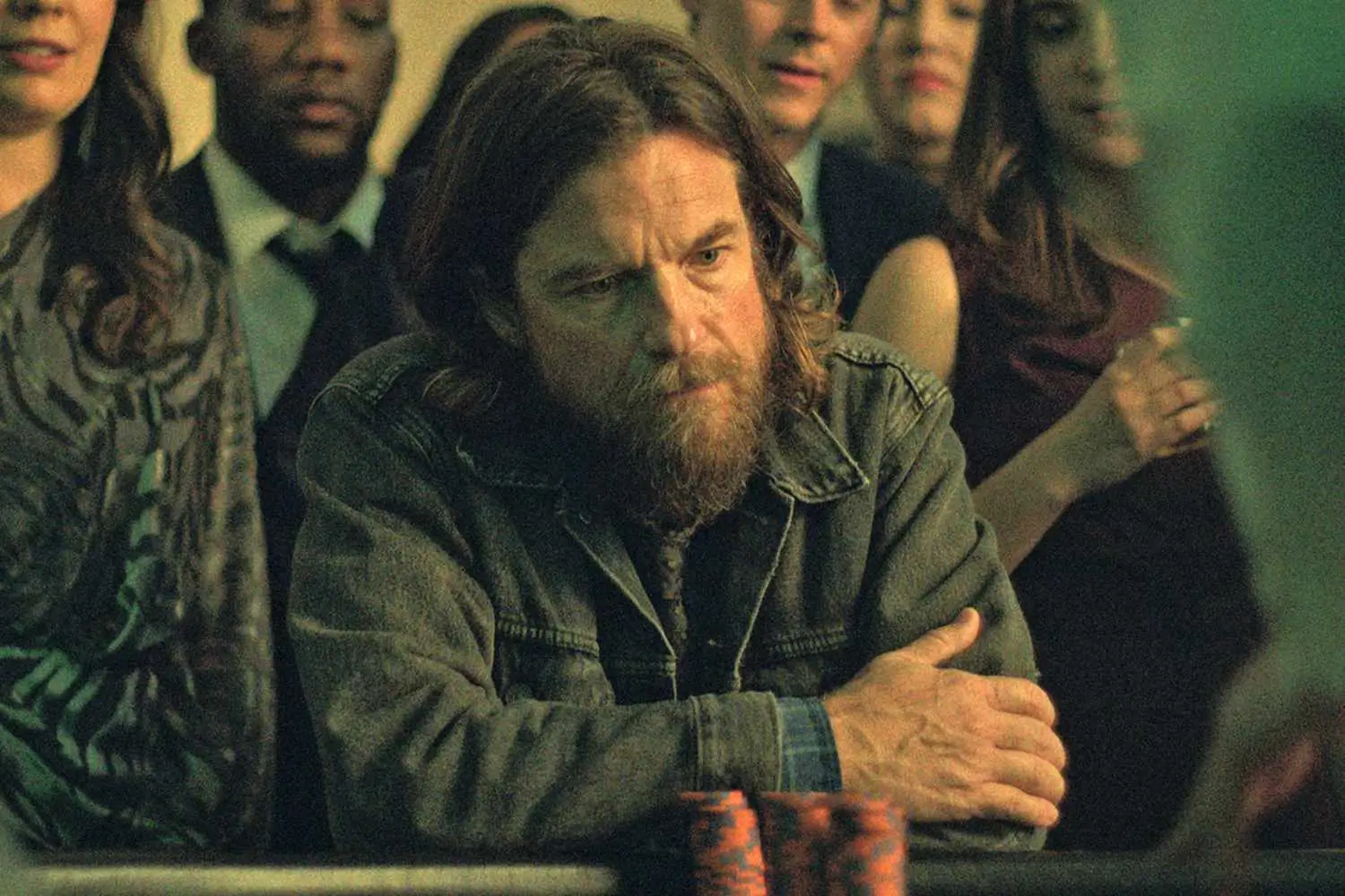 Cinematic scene featuring a long-haired bearded man leaning forward at a gaming table with arms crossed, wearing a faded dark jacket and an intense, focused expression. A softly blurred crowd gathers behind him under moody green-tinted lighting, creating a tense and intimate atmosphere