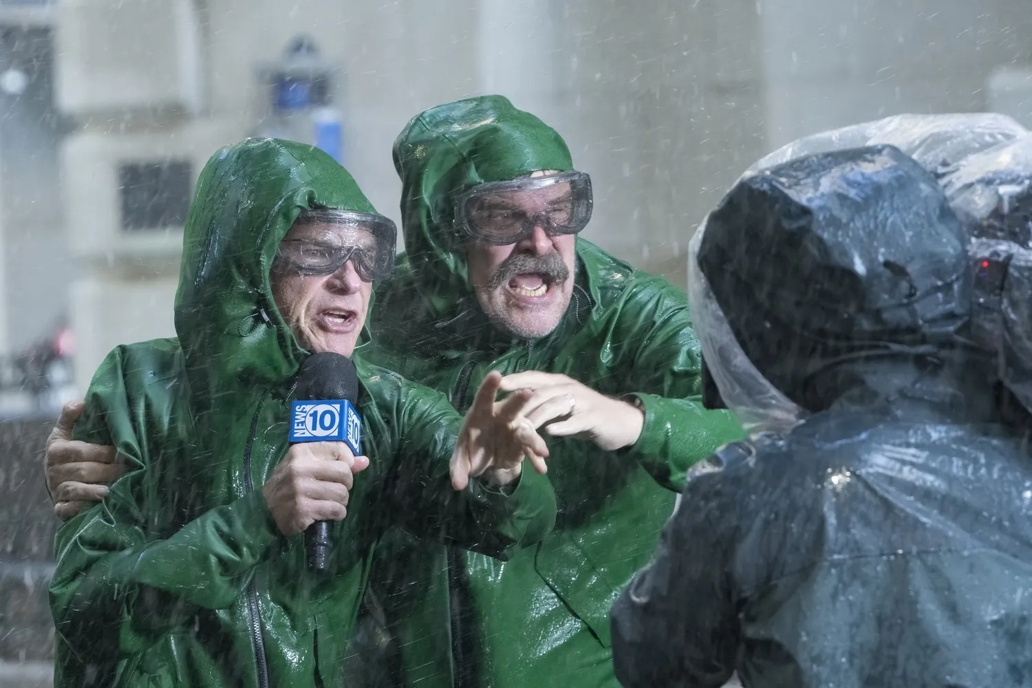 Behind-the-scenes style scene showing two men in green rain jackets and protective goggles reporting during heavy rain while facing a camera crew. One holds a news microphone as the other gestures forward, creating a chaotic, storm-chasing atmosphere with dramatic weather effects and broadcast production energy