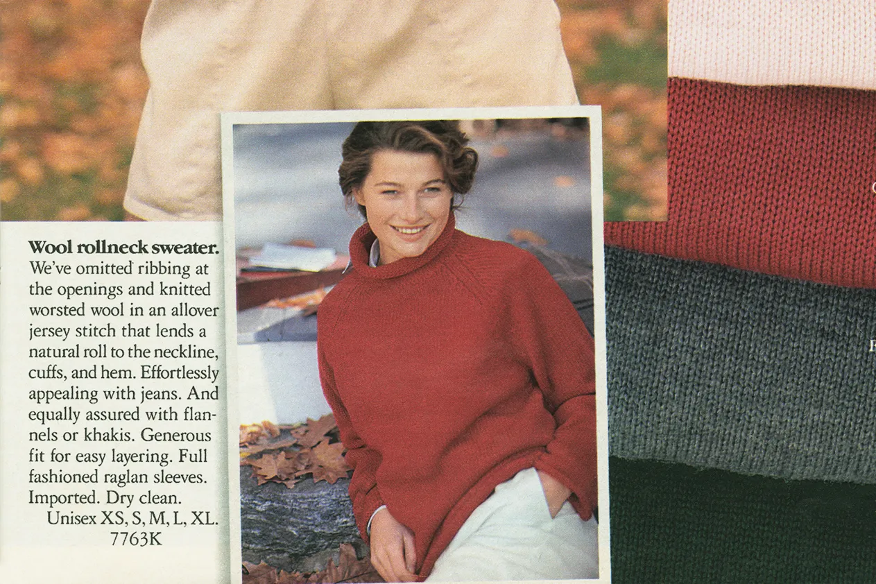 Vintage J.Crew catalog page featuring a model seated outdoors in autumn leaves while wearing a deep red wool rollneck sweater with relaxed raglan sleeves and soft rolled edges at the collar, cuffs, and hem. The knit’s oversized silhouette reflects the understated luxury and effortless layering central to late-1980s and early-1990s J.Crew styling. Alongside the image, editorial catalog copy describes the sweater’s worsted wool construction, natural rolled finish, and unisex fit, reinforcing the brand’s classic blend of functional American sportswear and refined seasonal dressing