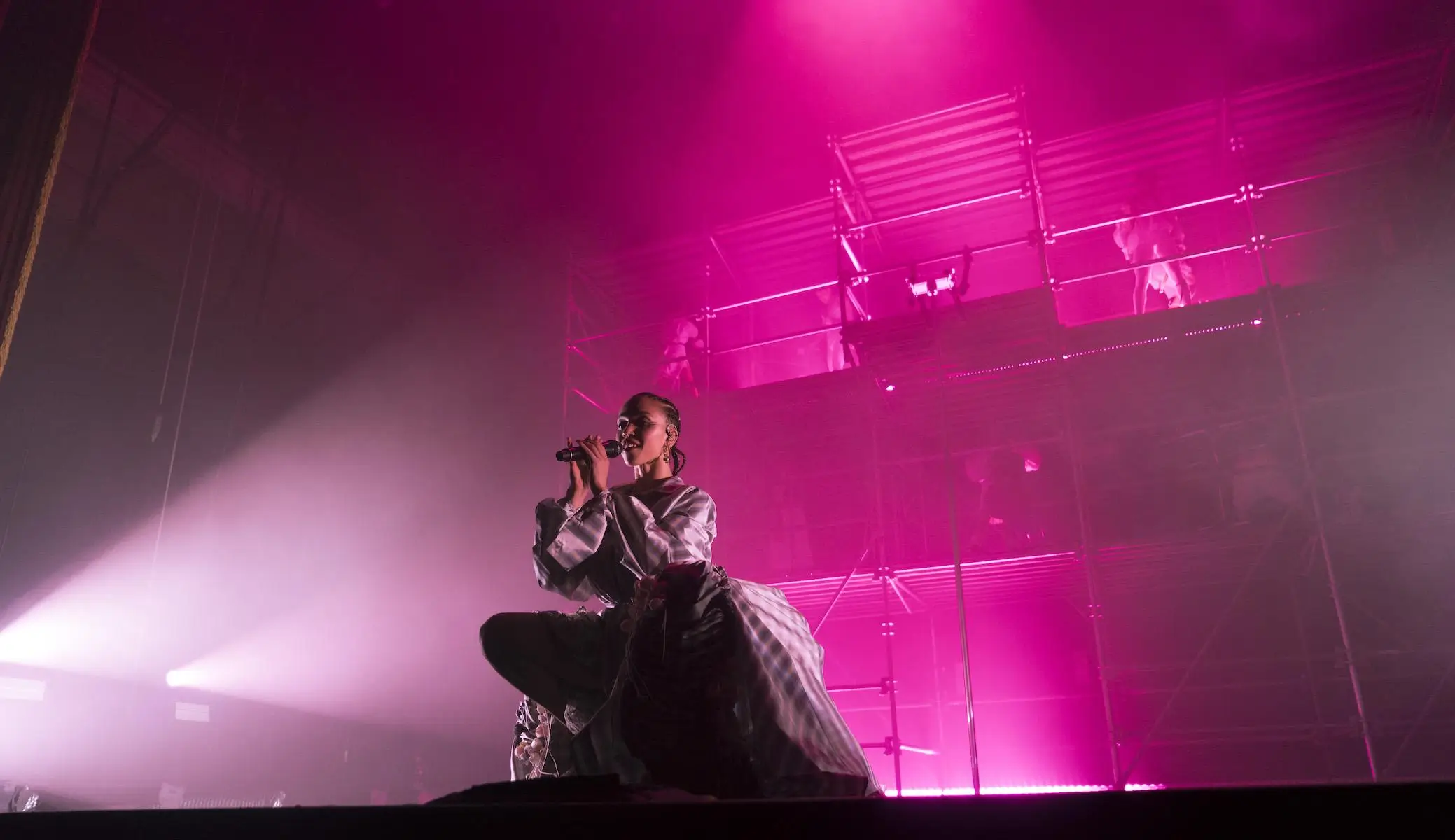 Concert performance photograph of a singer kneeling onstage beneath vivid magenta lighting, holding a microphone while framed by towering industrial scaffolding and atmospheric haze, creating a cinematic live-show atmosphere with dramatic shadows, layered staging, and high-energy arena production design