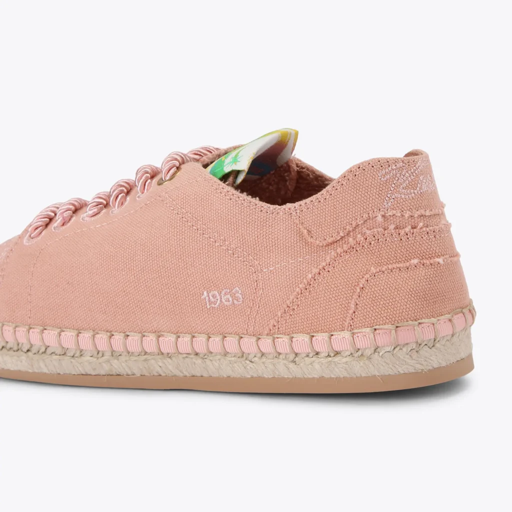 Side profile view of a pale pink canvas espadrille shoe featuring twisted rope laces through metal eyelets, tonal stitched paneling and embroidered heel detailing, braided jute trim around the midsole, rubber outsole, and subtle “1963” embroidery along the sidewall against a light gray background