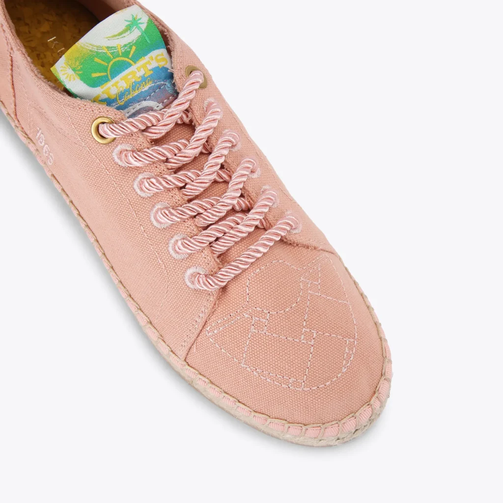 Close-up angled view of a pale pink canvas espadrille shoe featuring thick twisted rope laces threaded through tonal embroidered eyelets and gold-tone top eyelets, tonal geometric stitching across the toe box, braided jute trim around the sole, and a colorful tropical tongue label with sun and palm tree graphics