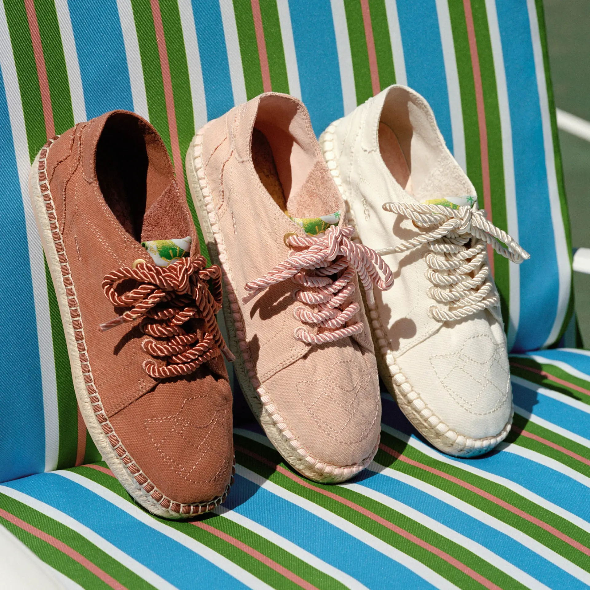 Three canvas espadrille shoe in brown, blush pink, and cream arranged on a striped outdoor lounge chair, each featuring thick twisted rope laces, tonal stitched geometric embroidery on the toe box, and braided jute midsoles in a sunlit resort-inspired setting