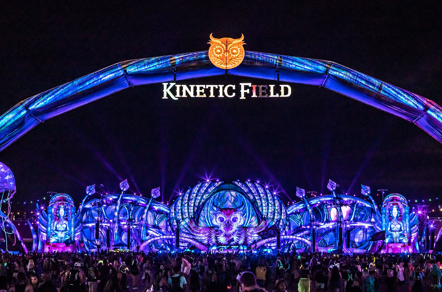 The towering kineticFIELD stage glows beneath the Las Vegas night sky at EDC, framed by a massive illuminated arch crowned with the festival’s iconic owl emblem. Bathed in electric shades of violet, blue, and magenta, the sprawling stage design blends futuristic fantasy with immersive rave spectacle as thousands of attendees gather below amid lasers, towering LED sculptures, and surreal architectural forms that transform the Speedway into a temporary neon metropolis