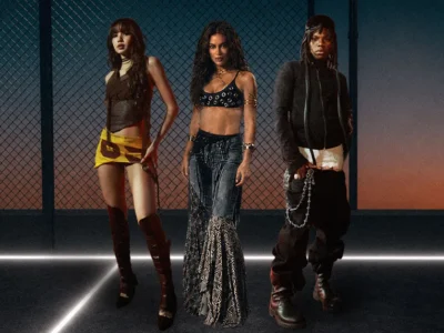 Alt text: Promotional image featuring three music artists standing in a stylized, stadium-like setting at dusk. From left to right: one wears a layered dark top with a yellow asymmetrical mini skirt and knee-high boots; the center figure wears a black embellished crop top paired with a textured floor-length skirt and metallic accessories; the right figure appears in layered black streetwear with chain details and oversized boots. The scene is framed by glowing floor lines and a chain-link backdrop, emphasizing a sleek, high-energy visual aesthetic tied to a global music campaign