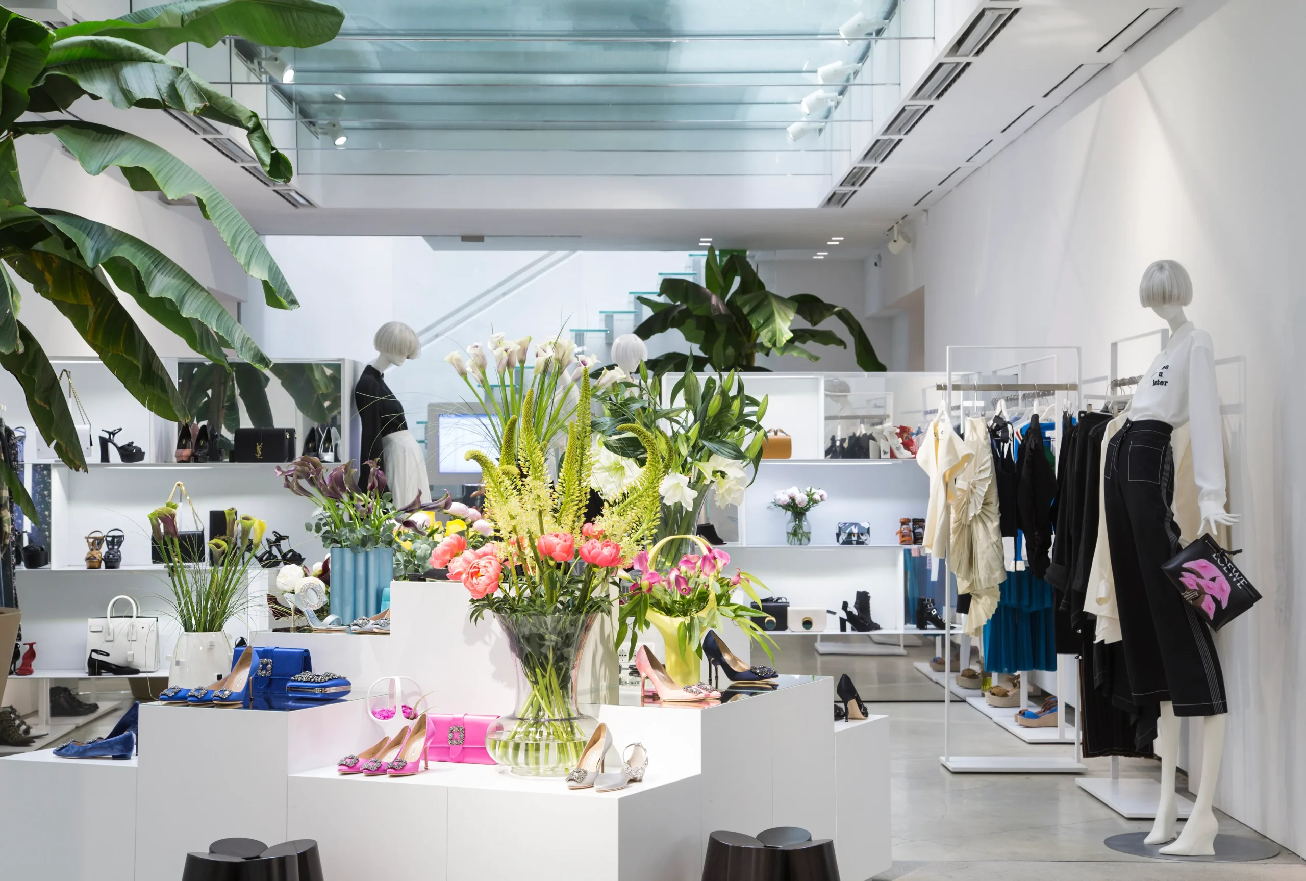 The bright interior of LuisaViaRoma in Florence features minimalist white architecture, designer footwear and apparel displays, tropical greenery, and floral installations, reflecting the retailer’s luxury fashion atmosphere during Pitti Uomo 2026