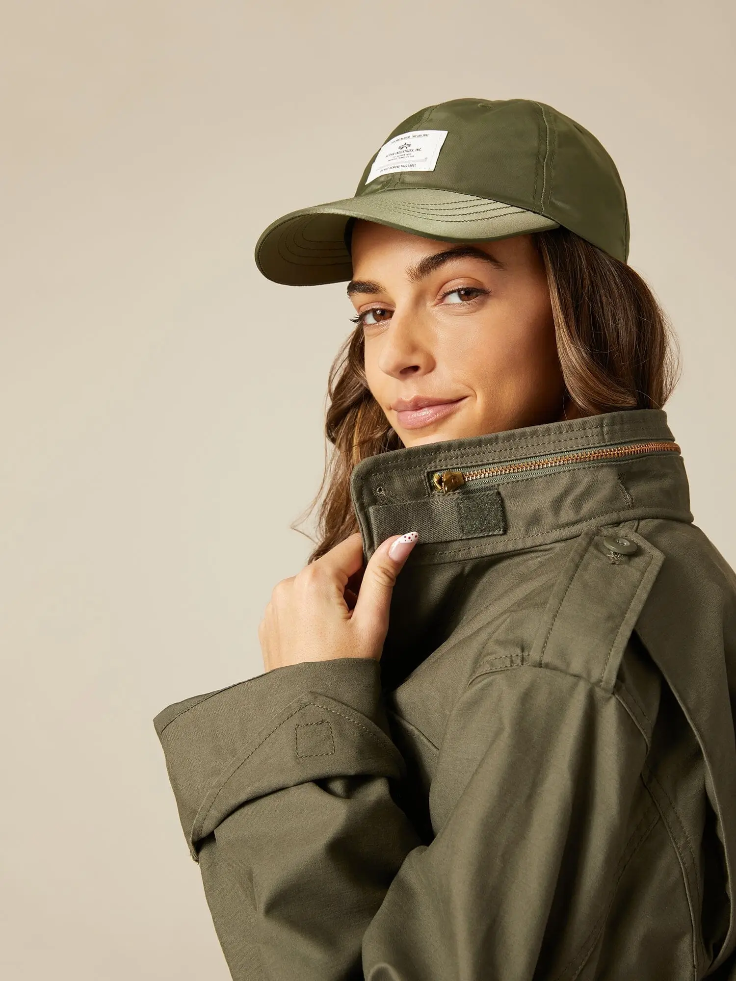 Editorial portrait showing a model wearing an olive green military-inspired field jacket styled with the stand collar fully raised and partially zipped to reveal the concealed hood compartment detail. Paired with a matching olive cap, the look highlights utility-inspired construction including epaulettes, adjustable cuff tabs, and structured outerwear elements against a clean neutral studio background