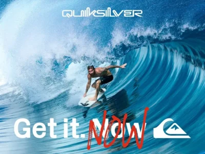 A dynamic promotional image from Quiksilver featuring a surfer carving through the hollow interior of a massive turquoise barrel wave. The cinematic ocean spray and deep-blue water textures create a high-energy action-sports atmosphere, while the bold “Get it. Now” typography and Quiksilver logo reinforce the campaign’s urgent, youth-driven surf identity