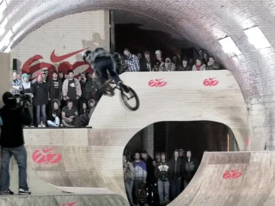 A BMX rider launches into a high-speed wallride inside Nike’s legendary 6.0 Tunnel Jam course, surrounded by spectators packed against towering wooden ramps beneath the curved brick ceiling of the abandoned Privett Tunnel in Hampshire, England. The underground skatepark’s raw industrial atmosphere, glowing tunnel lights, and bold Nike 6.0 branding amplify the intensity and experimental spirit of the iconic 2009 event