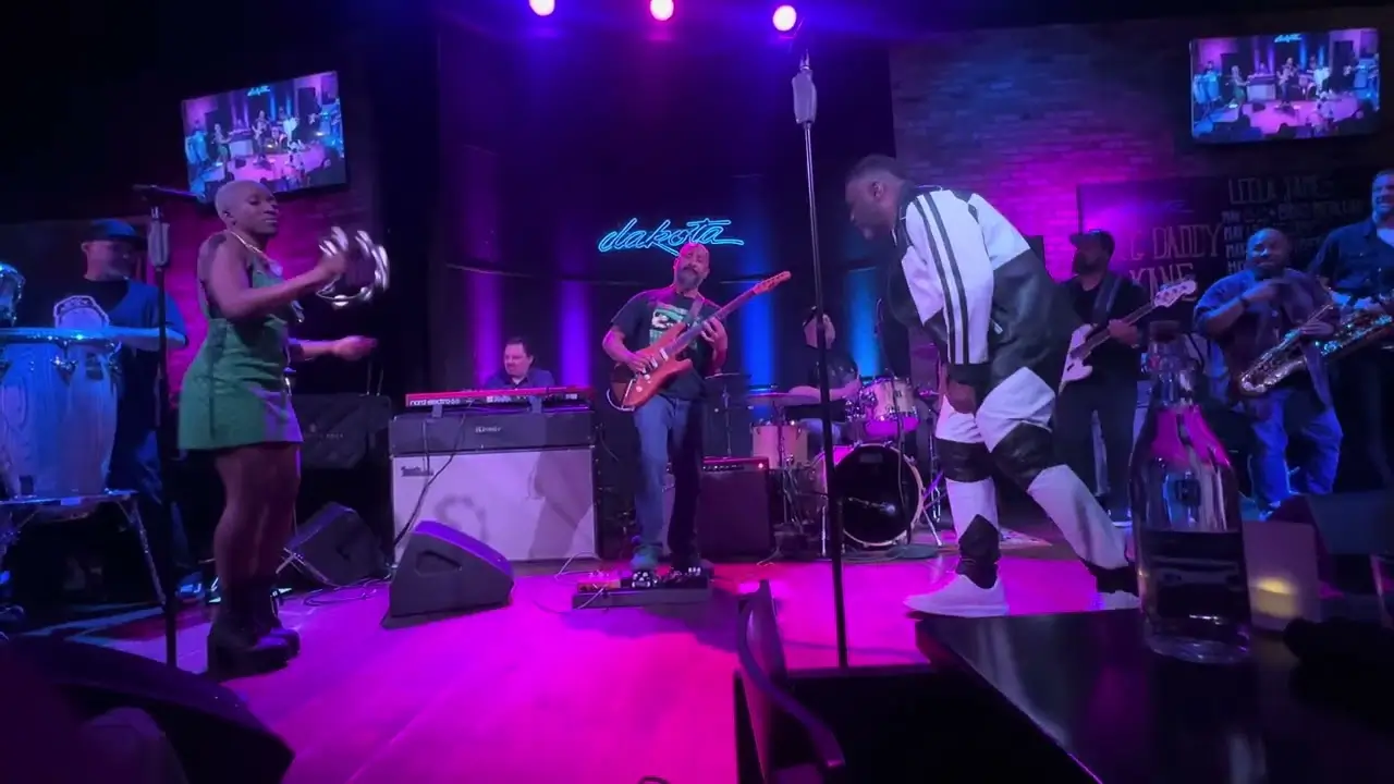 Live performance scene inside an intimate music venue illuminated with purple and blue stage lighting. A vocalist in a black-and-white performance outfit moves across the stage while interacting with a percussionist holding a tambourine. Behind them, a full band performs with guitar, keyboards, drums, bass, and saxophone, creating an energetic concert atmosphere with neon venue signage glowing in the background