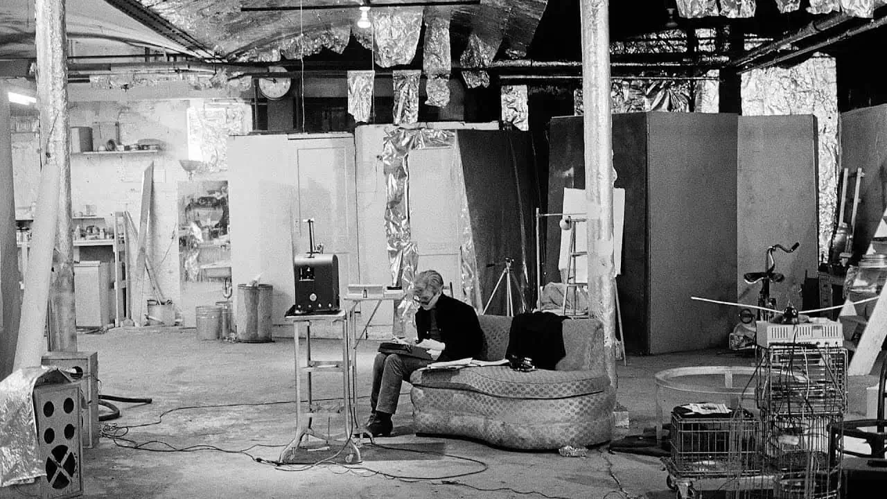 A stark black-and-white interior photograph capturing the raw industrial atmosphere of an artist’s studio space, filled with improvised structures, exposed wiring, metallic insulation, and scattered production equipment. At the center of the composition, a solitary seated figure leans forward on a worn couch, absorbed in reading or writing beneath harsh overhead lighting. Surrounding them are partition walls, carts, cables, cameras, and makeshift workstations, creating an environment that feels simultaneously abandoned, experimental, and intensely active beneath the surface.The reflective foil lining stretched across ceilings, columns, and walls gives the room an almost cinematic coldness, transforming the studio into something between a laboratory, underground film set, and conceptual art installation. The expansive negative space and visible infrastructural details emphasize process over polish, echoing the utilitarian aesthetic associated with 1960s avant-garde production spaces such as Andy Warhol’s Factory. Rather than presenting creativity as glamorous, the image foregrounds exhaustion, repetition, and the mechanical conditions behind artistic production, where silence, labor, and experimentation coexist inside an unfinished architectural shell