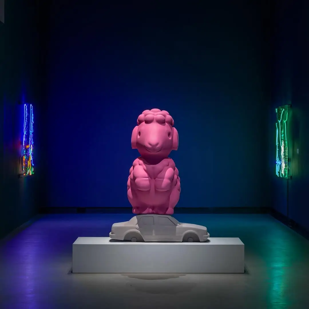 Gallery installation by Roda Medhat featuring a large pink sheep sculpture perched atop a white automobile, surrounded by neon artworks that explore themes of migration, identity, folklore, and contemporary Kurdish culture