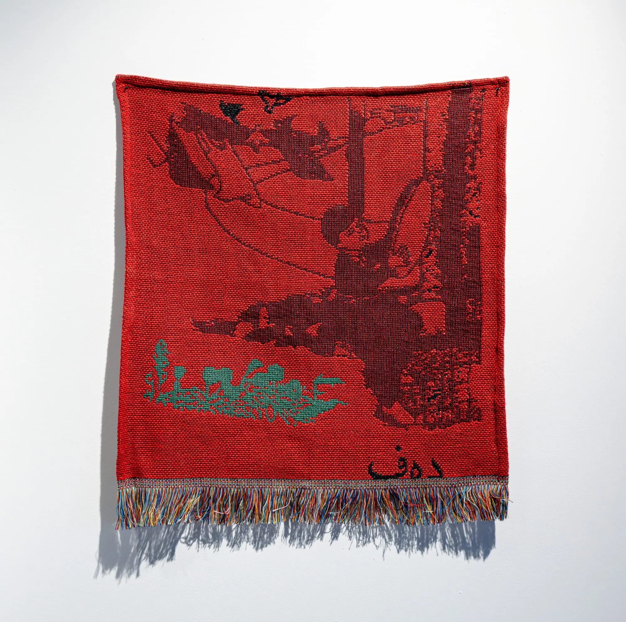 Handwoven red textile artwork by Roda Medhat depicting a monochromatic narrative scene with figures and landscape elements, finished with multicolored fringe and inspired by Kurdish storytelling tradition