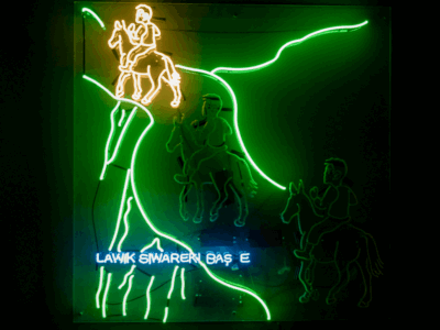 Animated neon artwork by Roda Medhat depicting horseback riders traversing a mountainous Kurdish landscape. Bright green LED outlines form winding cliffs and terrain, while glowing yellow and white figures on horseback illuminate the scene. A blue Kurdish-language text element runs across the lower section, creating a vibrant fusion of folklore, storytelling, and contemporary light-based installation art against a dark gallery backdrop