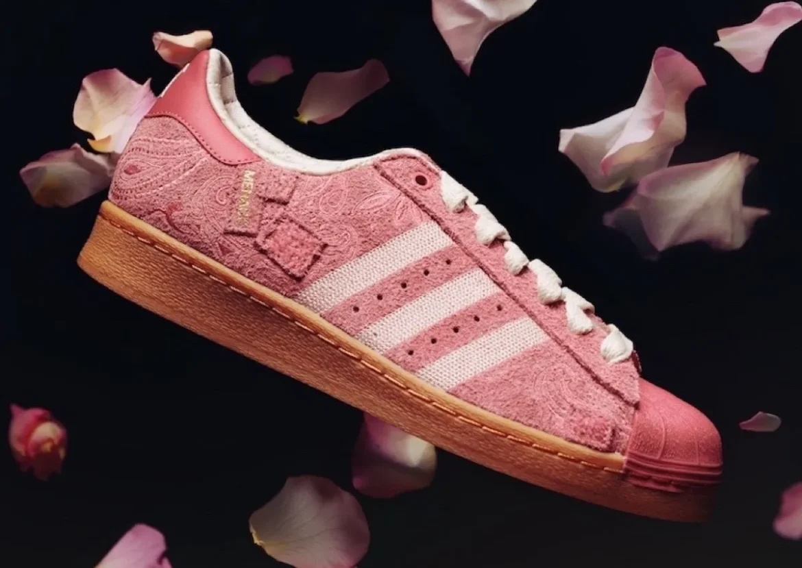 A soft pink Adidas Superstar shoe floats against a dark backdrop, surrounded by scattered rose petals that echo its color palette. The upper features a richly textured, floral-patterned fabric in varying shades of blush, complemented by the brand’s signature three white stripes along the side. A tonal pink rubber shell toe anchors the front, while a gum sole adds warmth and contrast beneath. Cream laces and a lightly padded collar soften the structure, giving the shoe a romantic, almost vintage feel that blends streetwear heritage with delicate, ornamental detail