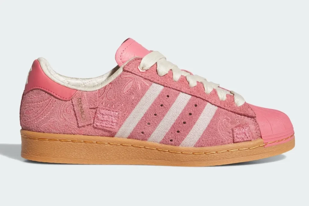 A pink Adidas Superstar shoe is presented in a clean side profile, highlighting its balanced proportions and material interplay. The upper is constructed from textured pink suede, subtly embossed with a paisley pattern that adds depth without overpowering the design. The signature three stripes appear in off-white woven fabric, paired with matching cream laces and a softly padded, satin-like inner lining for a refined contrast