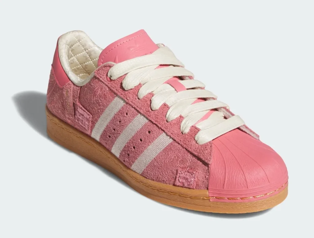 A pink Adidas Superstar shoe is shown from a front three-quarter angle, highlighting both its structure and layered textures. The upper is built from soft, suede-like material with subtle paisley-style embossing, giving the surface a tactile, decorative depth. The signature three stripes appear in off-white, coordinating with the thick cream laces that sit prominently over a tonal pink tongue. Up front, the glossy rubber shell toe in matching pink adds a sculpted contrast to the matte upper. Inside, a quilted, satin-like lining peeks through, introducing a refined, almost luxe interior detail. The gum rubber sole grounds the design with a warm, classic finish, balancing the soft pink palette with a heritage touch