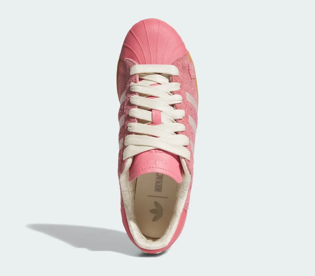 A pink Adidas Superstar shoe is shown from a top-down perspective, centering the lace structure and toe design. The wide cream laces run neatly over a tonal pink tongue, creating a soft contrast against the textured suede upper. Subtle paisley-style embossing is visible across the pink surface, adding depth without overwhelming the monochrome palette. At the front, the glossy pink rubber shell toe stands out with its ridged detailing, anchoring the silhouette. Inside, a quilted, satin-like lining in a light tone surrounds the collar, while the insole reveals the Adidas Trefoil branding, completing the composition with a clean, refined finish
