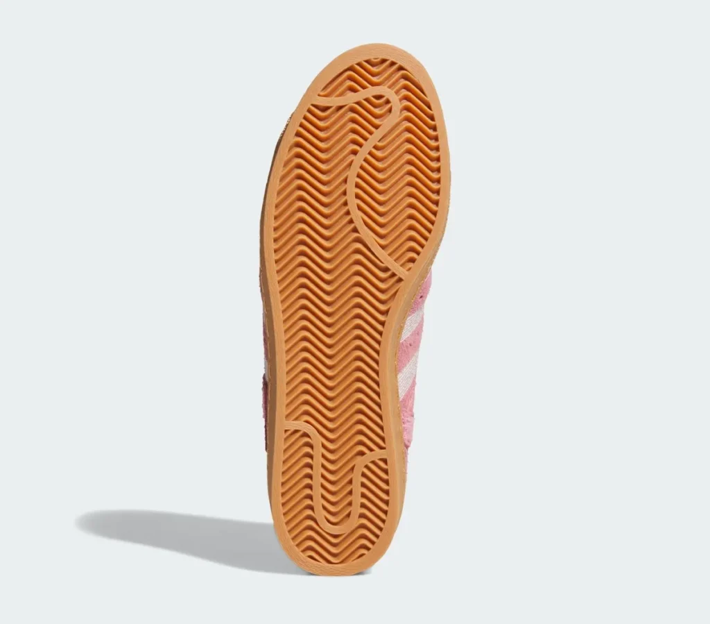 The outsole of the Adidas Superstar is shown in a full-length view, highlighting its classic gum rubber construction. A dense herringbone traction pattern runs across the entire sole, designed for grip and durability while maintaining flexibility. Subtle pivot points and curved grooves are integrated into the forefoot and heel areas, enhancing movement and control. The warm gum tone contrasts with the softer pink upper seen elsewhere, reinforcing the shoe’s heritage basketball roots while grounding the overall design with a functional, time-tested foundation