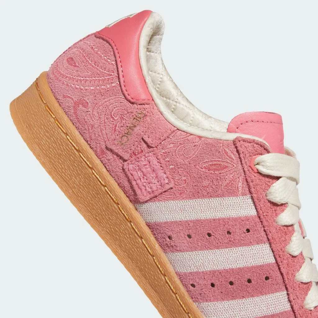 A close-up of the Adidas Superstar highlights the richness of its materials and detailing. The pink suede upper is densely textured with an embossed paisley pattern, giving the surface a layered, almost velvety depth. The signature three stripes appear in off-white woven fabric, contrasting subtly against the tonal pink base. Fine stitching and small perforations line the mid-panel, while a delicate gold script branding sits near the eyelets, adding a refined accent. The padded collar reveals a quilted, satin-like lining in a soft cream tone, enhancing the shoe’s comfort and elevated feel. Beneath, the gum rubber midsole—with visible stitching—grounds the composition, balancing the ornate upper with a durable, heritage-inspired foundation