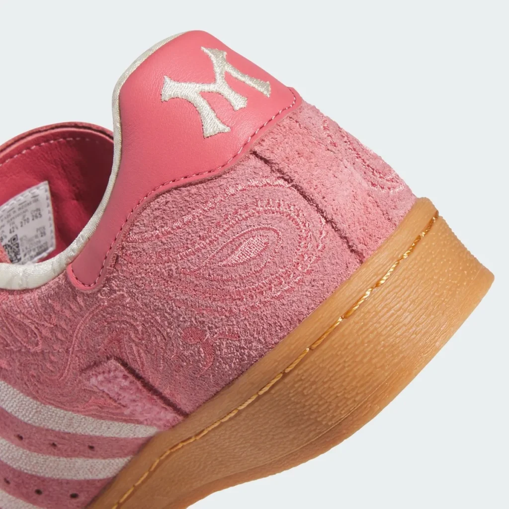 A close-up of the heel area of the Adidas Superstar emphasizes its layered textures and branding details. The pink suede upper features a finely embossed paisley pattern, giving the surface a soft, dimensional quality. At the top, a smooth pink leather heel tab is finished with a contrasting white embroidered monogram-style logo, standing out crisply against the tonal base. The transition between materials is defined by precise stitching, while the gum rubber midsole below adds a warm, textured foundation. The combination of plush suede, clean leather, and classic sole construction reinforces the shoe’s blend of ornamental design and heritage structure