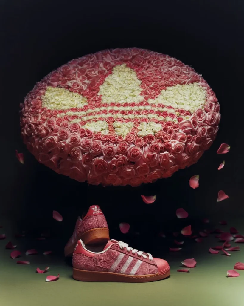 A pink Adidas Superstar shoe sits in the foreground beneath a dramatic, floating installation shaped like the Trefoil logo, constructed entirely from tightly packed roses. The floral structure blends soft pink and cream tones to form the iconic three-leaf motif, suspended against a deep, shadowy backdrop. Scattered petals drift through the scene, reinforcing the romantic, almost surreal atmosphere. Below, the sneaker mirrors the installation’s palette: a textured pink suede upper with subtle paisley embossing, off-white three stripes, cream laces, and a tonal pink shell toe, all grounded by a warm gum sole. The composition connects product and environment, turning the shoe into an extension of the floral sculpture above—heritage sportswear reframed through a lush, ornamental lens