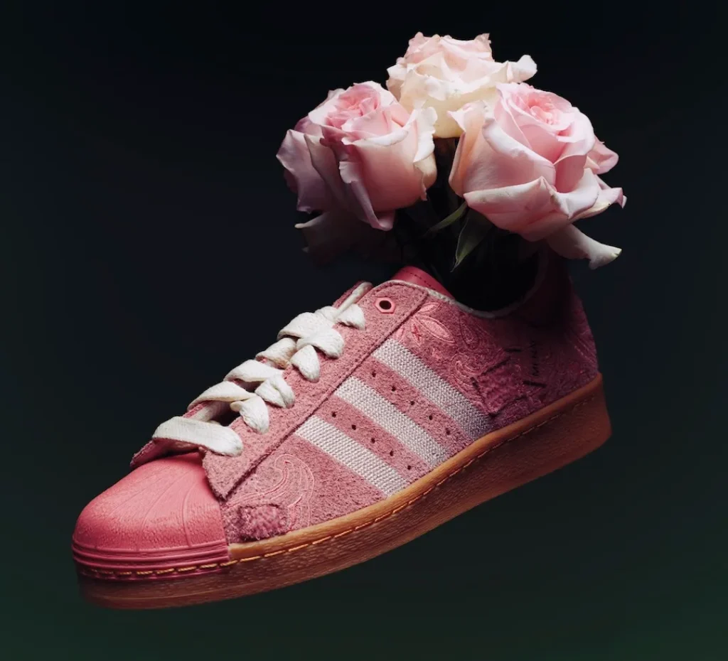 A pink Adidas Superstar shoe appears suspended in a dark, atmospheric setting, with a bouquet of soft pink roses blooming from its opening. The composition merges product design with floral still-life, creating a romantic, almost surreal visual. The shoe’s upper is crafted from textured pink suede, detailed with subtle paisley embossing that echoes the organic curves of the petals above. Off-white three stripes and thick cream laces introduce gentle contrast, while the tonal pink shell toe adds a sculptural, glossy finish at the front. Beneath, the gum rubber sole grounds the design with a warm, heritage-inspired base. The roses—delicately layered and softly lit—mirror the sneaker’s palette, reinforcing a cohesive, ornamental narrative where classic sportswear is reimagined through a lush, floral lens