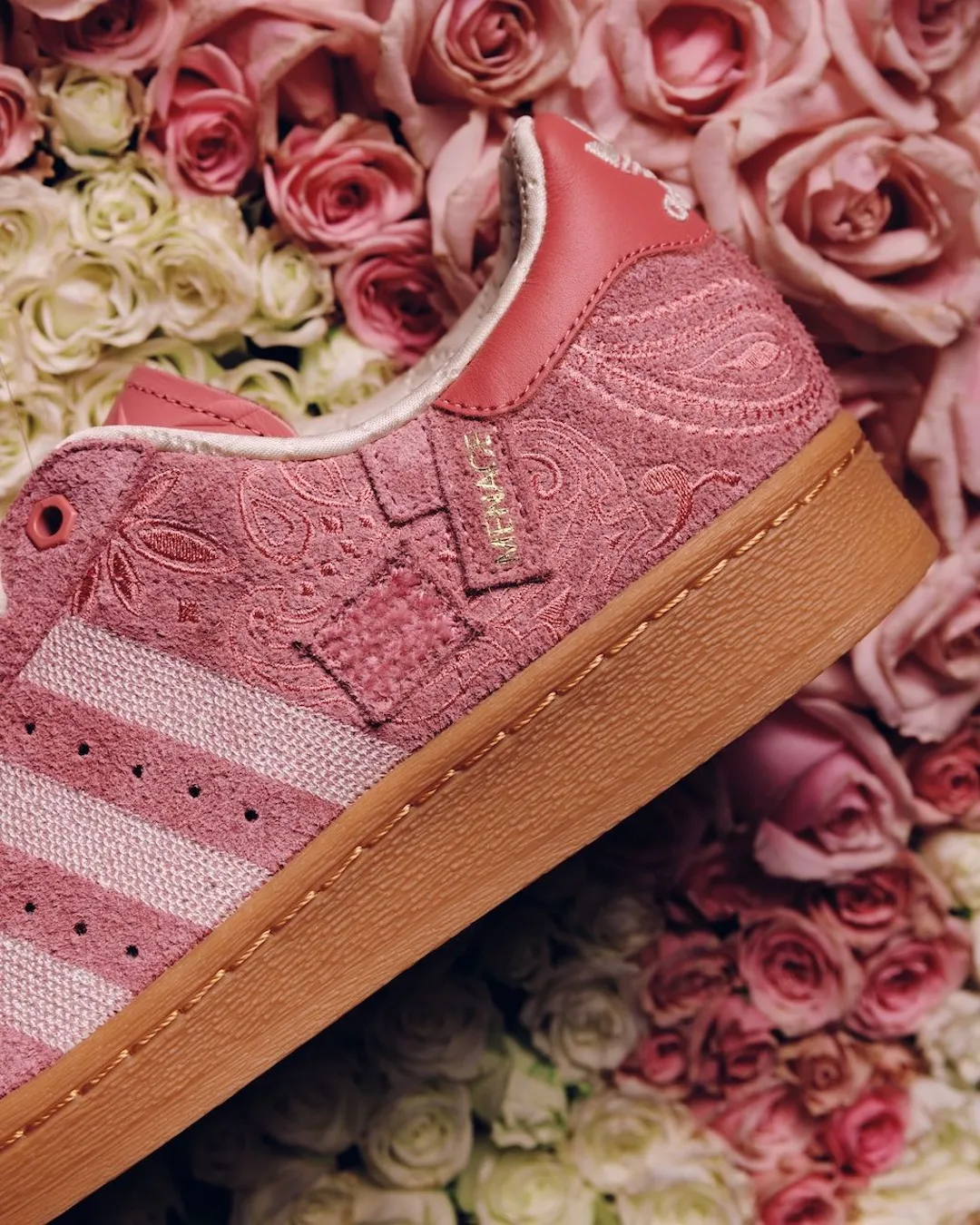 A close-up of the Adidas Superstar is set against a dense backdrop of blooming roses, blending product and environment into a single tonal composition. The pink suede upper, richly embossed with a paisley pattern, mirrors the organic curves and textures of the surrounding petals. Off-white woven three stripes cut through the surface with subtle contrast, while fine perforations and stitching add structure to the design. A small gold script detail near the eyestay introduces a refined accent, catching light against the matte suede. The padded collar reveals a soft, satin-like lining in a pale tone, enhancing the sense of comfort and finish. Beneath, the gum rubber sole—with visible stitching—anchors the composition, its warm hue complementing the layered pinks and creams of both the sneaker and the floral backdrop