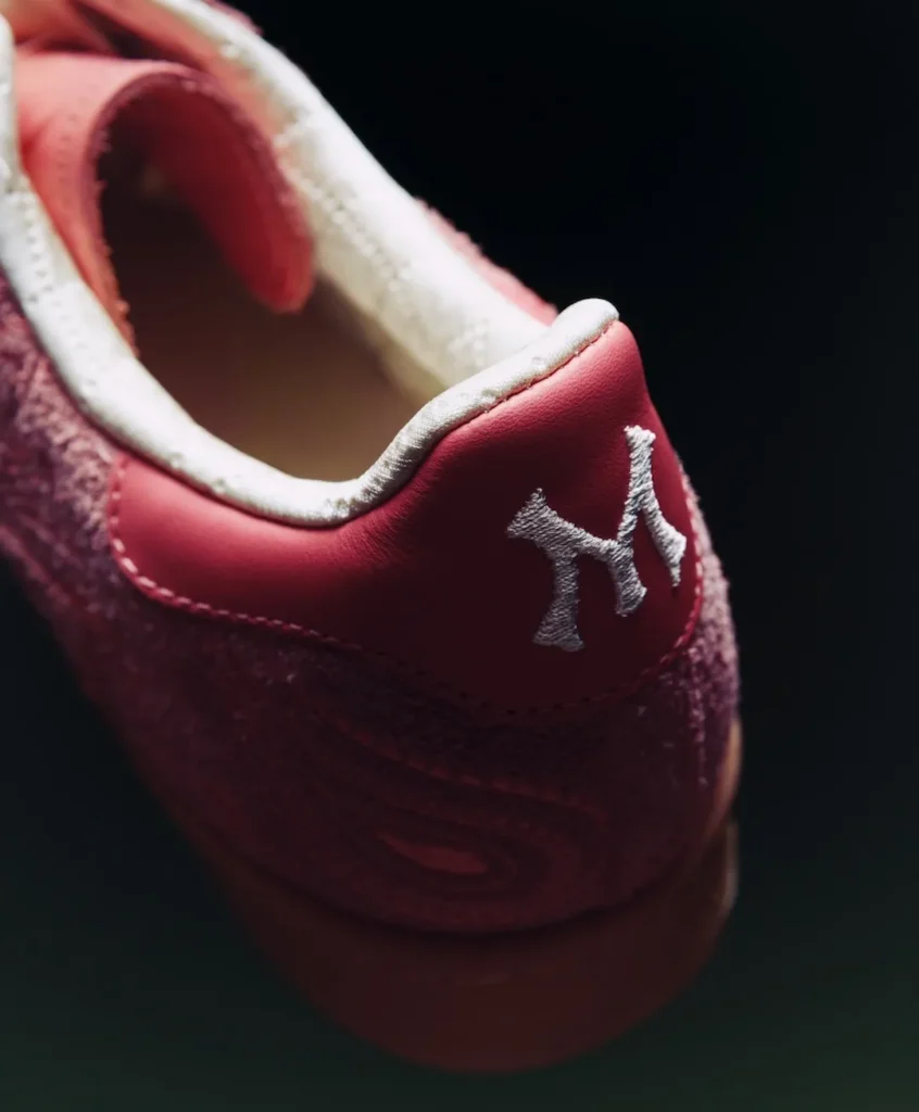 A close-up of the heel area reveals the Adidas Superstar’s refined finishing and material contrast. The smooth pink leather heel tab is sharply defined, carrying a white embroidered monogram-style logo that stands out crisply against the saturated tone. Surrounding it, the suede upper shows a subtle paisley embossing, its texture catching light in soft variations. The collar is lined with a quilted, satin-like material in a pale cream shade, adding a plush, almost luxurious interior feel. Fine stitching traces the edges with precision, while the gum rubber sole below provides a warm, grounded base. The tight framing emphasizes craftsmanship—where heritage structure meets delicate, ornamental detailing