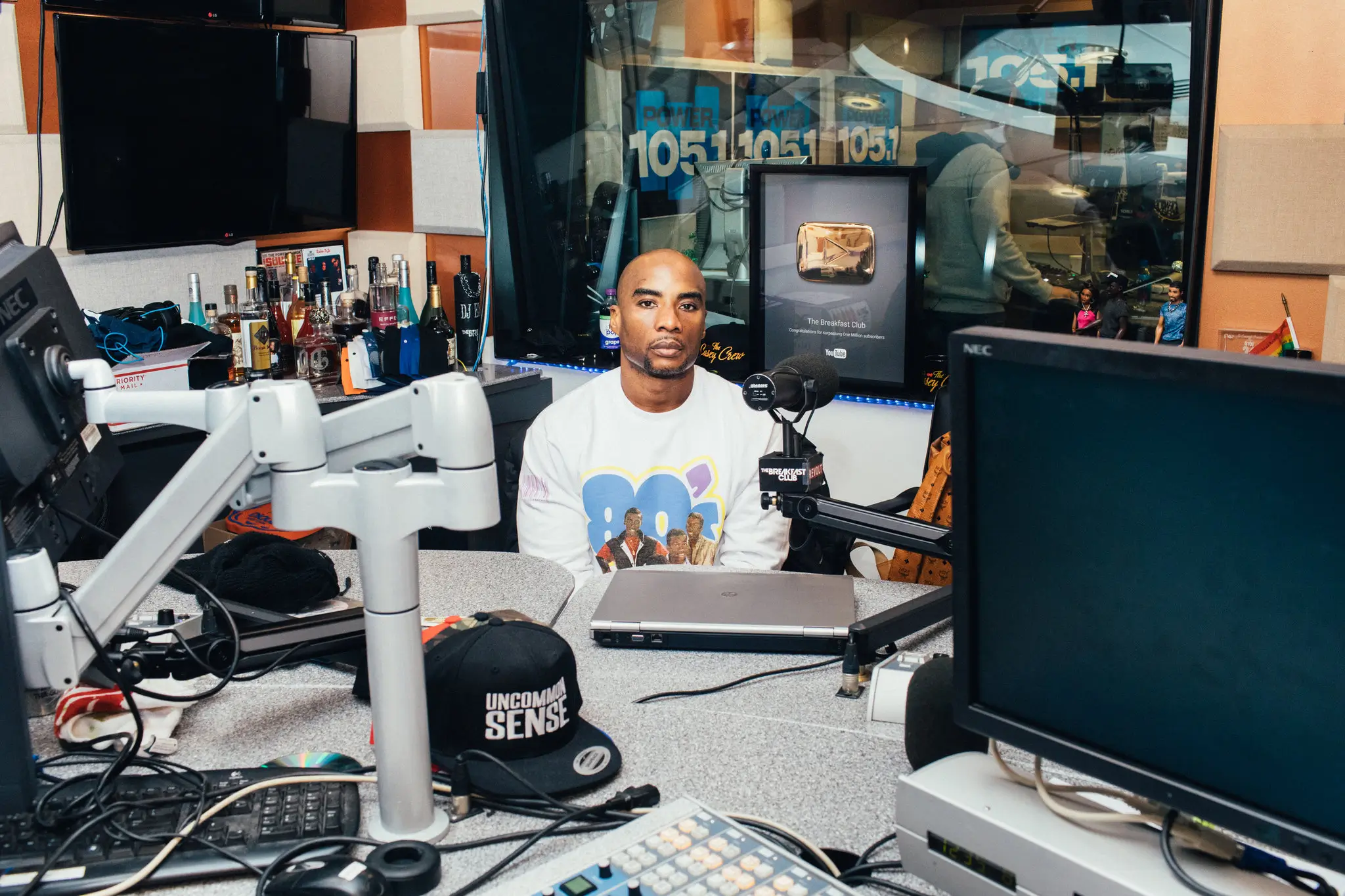 A portrait inside The Breakfast Club radio studio at Power 105.1 featuring a host seated behind the broadcast desk, surrounded by suspended microphones, monitor arms, computer screens, audio equipment, and studio clutter. Behind him, a framed YouTube Creator Award and Power 105.1 branding are visible through the glass into the production booth, emphasizing the show’s long-running presence across radio and digital media