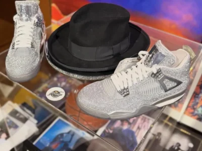 Display of the unreleased Michael Jackson Air Jordan 4 “Glitter Glove” Sample featuring two silver glitter-covered sneakers arranged beside a black fedora hat inside a transparent display case. Chrome accents, white laces, and shimmering textures reference Michael Jackson’s iconic rhinestone glove, while surrounding memorabilia reinforces the tribute to the King of Pop’s stage legacy
