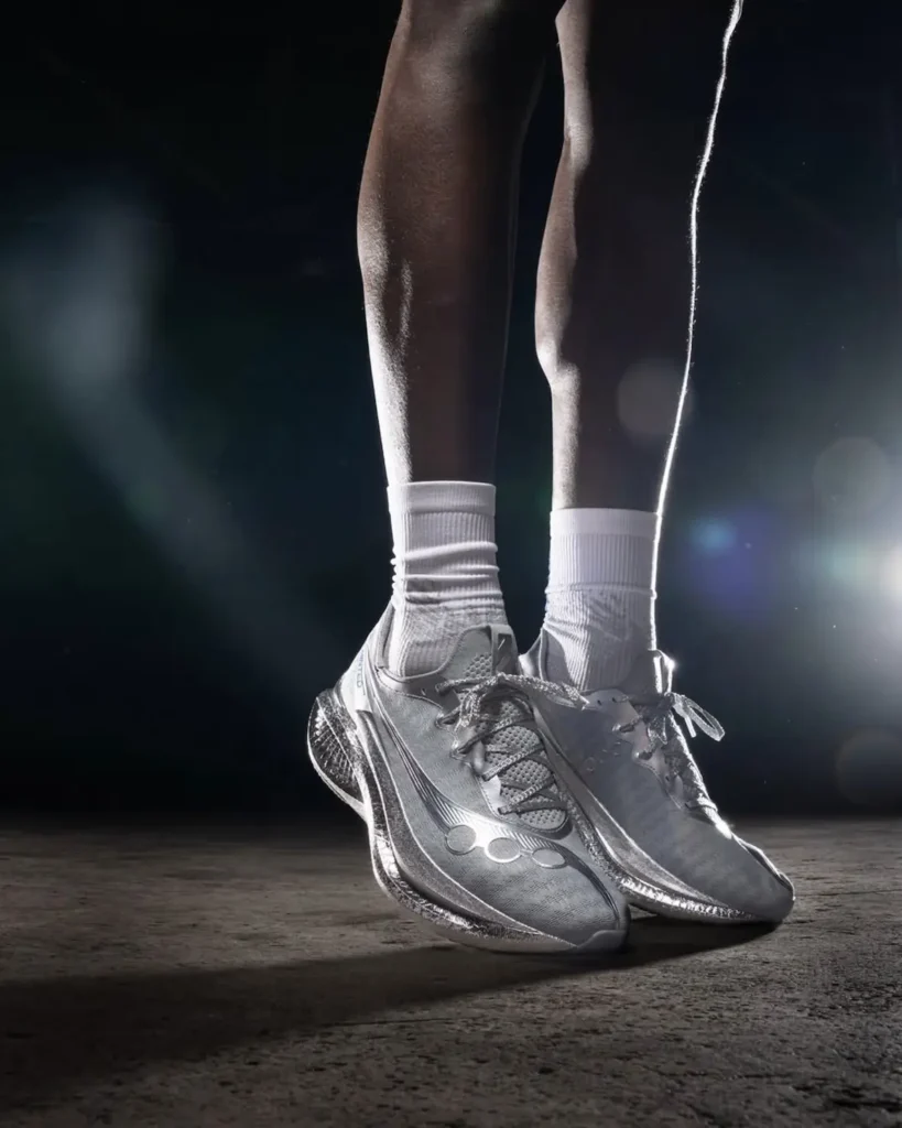 A close-up frames a runner’s lower legs and feet standing on a dimly lit surface, wearing white crew socks and sleek metallic running shoes. The shoes feature a smooth, reflective upper with sculpted contours and a textured, performance-focused sole. Bright, directional lighting creates lens flares and highlights along the edges, emphasizing the shoe’s shine and the definition of the calves against the dark, minimal background