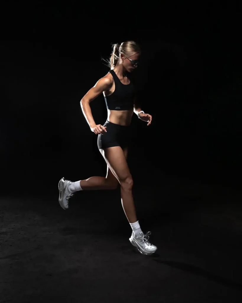 A runner moves through a dark, studio-lit space, captured mid-stride with one leg extended forward and the other lifting behind. They wear a black sports bra and fitted shorts, paired with white crew socks and metallic running shoes that catch the light with a subtle sheen. The focused lighting sculpts the body’s movement and highlights the reflective detailing of the footwear, while the black background isolates the figure, emphasizing rhythm, control, and forward motion